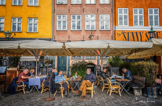A fine art photograph of people sitting at an out door restaurant
in the old port of Nyhavn in Copenhagen with brightly coloured rowhouses behind them